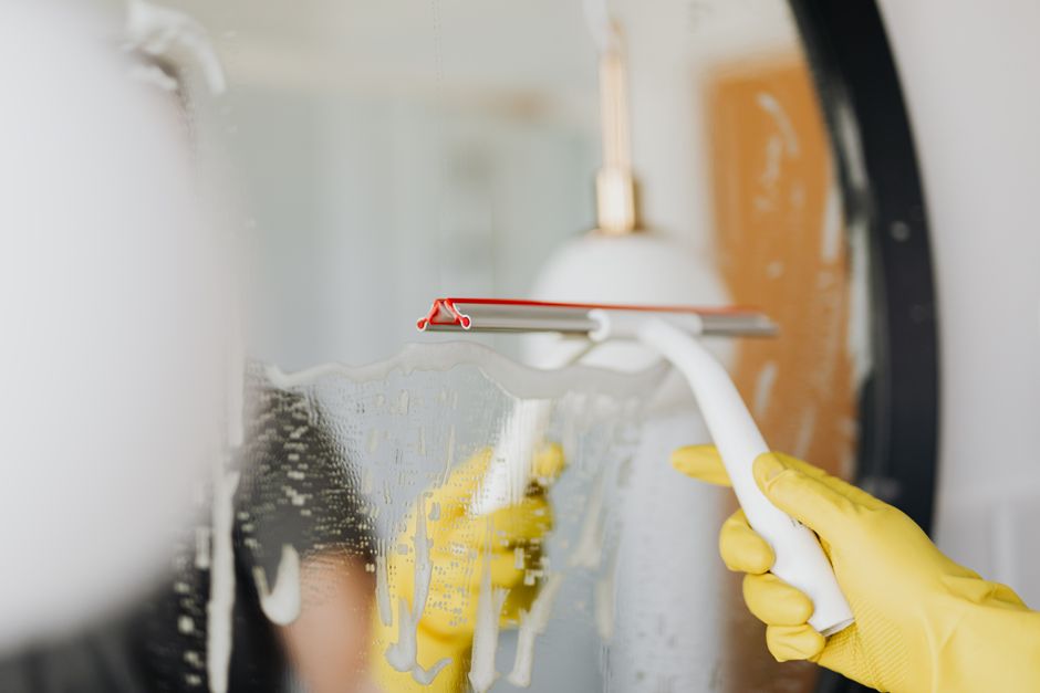 Anonymous person in yellow rubber gloves wiping water by squeegee from mirror in bathroom
